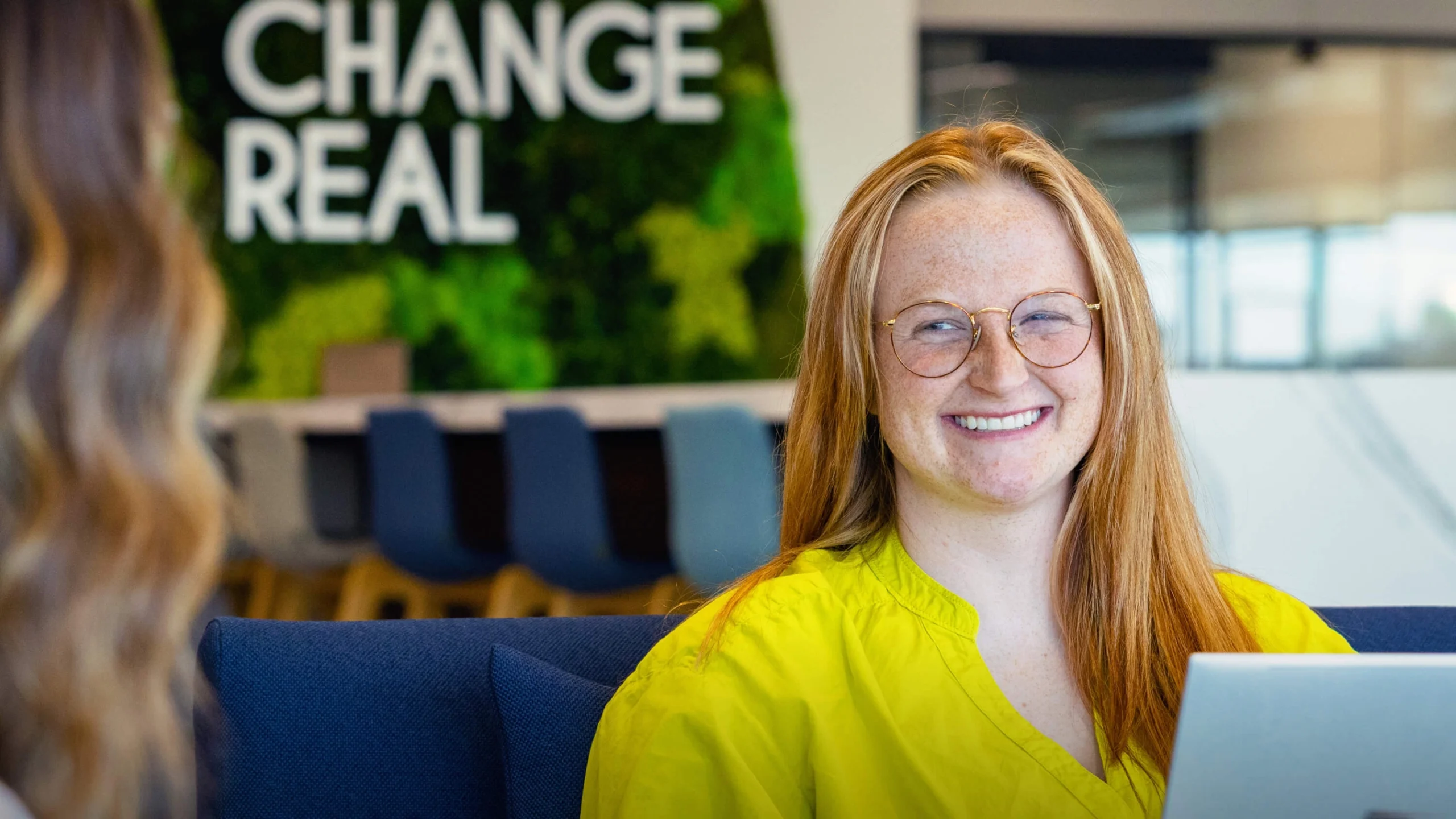 Female employee working on laptop in office with "Make Change Real" mission sign.