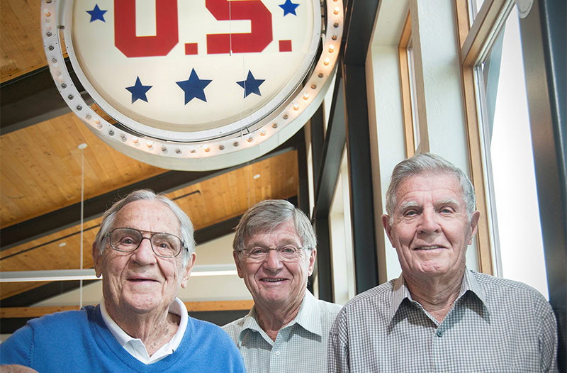 Three older men smiling under retro U.S. signage